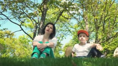 Anne ve oğlu mesafe içine bakarak ve yeşil çimenlerin üzerinde parkta otururken konuşurken aile tatili