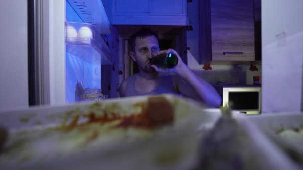 男人在冰箱里找食物 一个没有刮胡子的男人手里拿着一瓶啤酒 他在找冰箱里可以吃的东西 夜间饥饿 学士的冰箱 满是陈腐的食物 — 图库视频影像