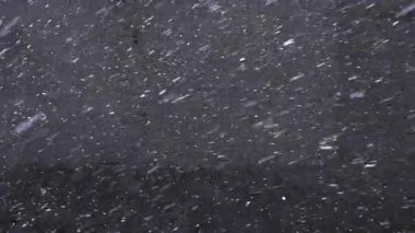 Kar taneleri gri asfalta düşer, manzaranın üstüne. İlk kar yağışı. Kaldırıma kar yağar. Ağır çekim. Kış havası. Soyut doğal arkaplan, hareket halindeki beyaz parçacıklar. 