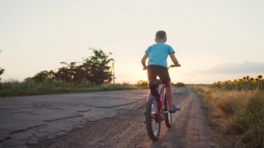 Bir çocuk kır yolunda bisiklete biner. Çocuk bisikleti. Aktif yaz tatili. Şehrin dışında bisikletiyle gezen mutlu çocuk. Bir çocuk ayçiçeği tarlasının yanında at sürüyor. Gün batımı. Sabah bisikleti.