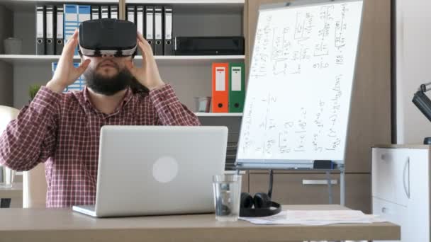 Homem barbudo em um escritório tira o fone de ouvido de realidade virtual VR da mesa e o coloca em sua cabeça — Vídeo de Stock