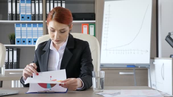 Hermosa mujer de negocios pelirroja en su escritorio en la oficina mirando a través del papeleo — Vídeo de stock