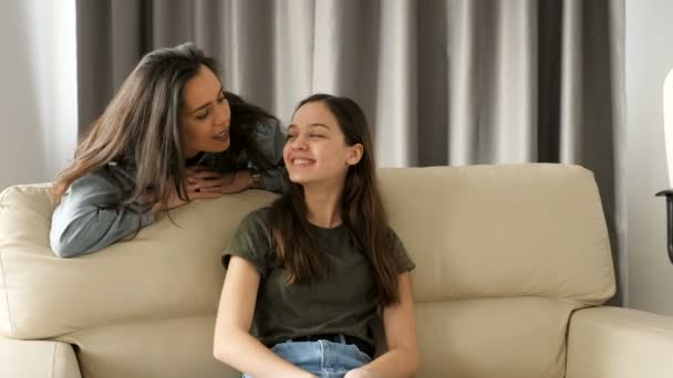 Младшая сестра сидит на диване, пока старшая щекочет ее — стоковое видео