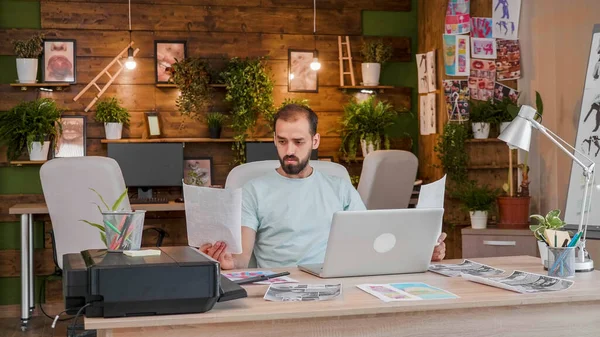 Jovem designer olhando para uma folha na mão enquanto trabalhava no laptop — Fotografia de Stock