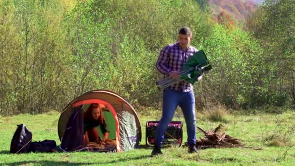 Pojkvännen sitter på campingplatsen medan flickvännen förbereder tältet — Stockvideo