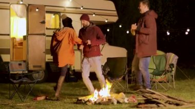 Soğuk bir sonbahar gecesinde kamp ateşi etrafında dans eden bir grup yakın arkadaş.
