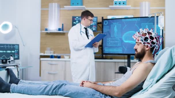 Doctor analysing patient sitting on bed and wearing brain scanning device — Stock Video