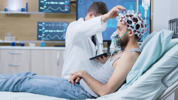 El médico sostiene la tableta y realiza ajustes en los sensores cerebrales que atraen al paciente — Vídeo de stock