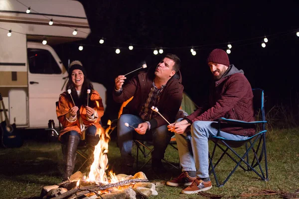 Hombre soplando un malvavisco caliente mientras sus amigos se ríen alrededor del fuego del campamento — Foto de Stock