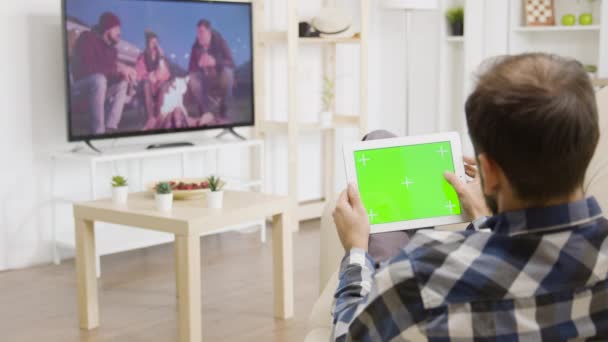 Mann berührt einen digitalen Tablet-PC mit grünem Bildschirm, während er auf den Fernseher schaut — Stockvideo
