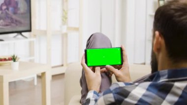 Beyaz adam televizyona bakarken elinde yeşil ekran akıllı telefon tutuyordu.