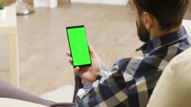 Kanepede oturan genç adam yeşil ekran telefonuna bakıyor.