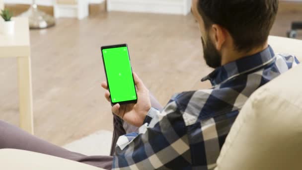 Jongeman zittend op de bank kijkend naar smartphone met groen scherm erop — Stockvideo