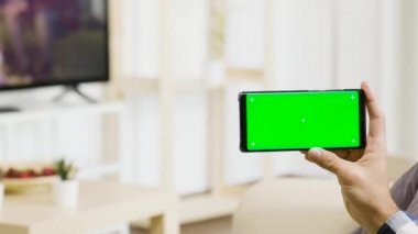 Modern ve parlak oturma odasında bir adam elinde yeşil bir ekran telefonu tutuyor.