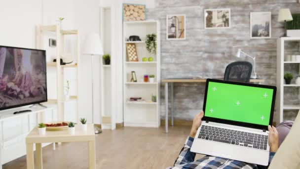 Man liggend op de bank in zijn woonkamer met een groene scherm laptop — Stockvideo