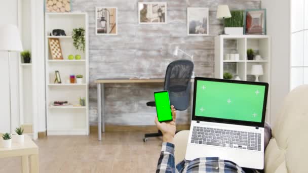 Hombre acostado en el sofá sosteniendo una computadora portátil de pantalla verde y un teléfono inteligente en sus manos — Vídeos de Stock