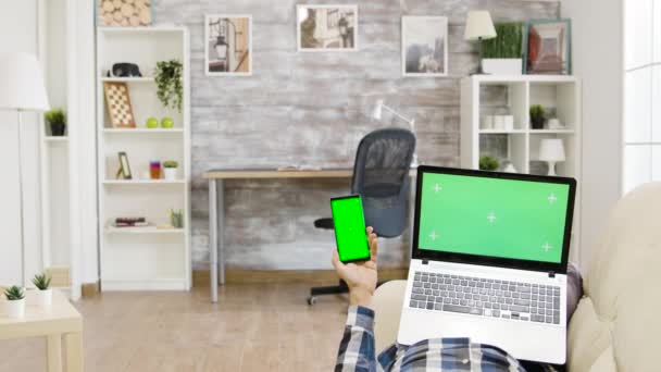 Man lying on the sofa holding a green screen laptop and smartphone in his hands — Stock Video