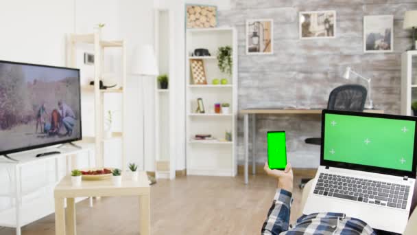 В яркой и хорошо освещенной гостиной мужчина держит ноутбук и телефон с зеленым экраном на них — стоковое видео