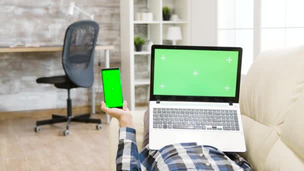 Tembakan Parallax seorang pria tergeletak di sofa, memegang layar hijau smartphone dan laptop di tangan — Stok Video