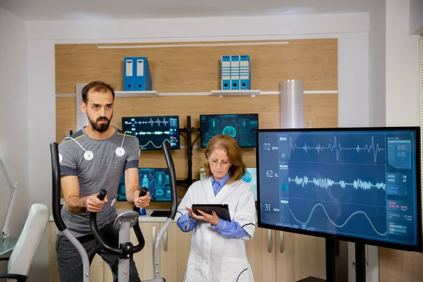 Atleta masculino que hace un esfuerzo físico en el escalón y tiene electrodos conectados a él y al médico que sigue la evolución de su tableta —  Fotos de Stock