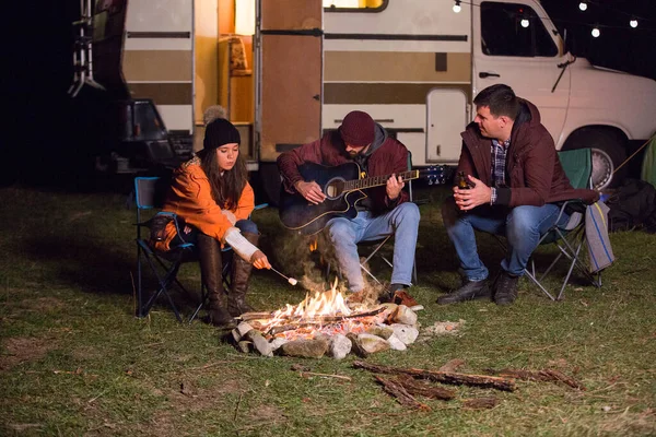 Hombre jugando en la guitarra alrededor de fuego de campamento mientras sus amigos ar tostado malvaviscos — Foto de Stock