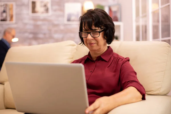 Old elderly woman on her sofa working on a modern laptop in her cozy living room — Stock Photo, Image