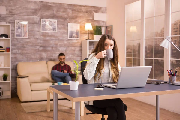 Kaukasische Frau genießt eine Tasse Kaffee, während sie von zu Hause aus am Laptop arbeitet — Stockfoto