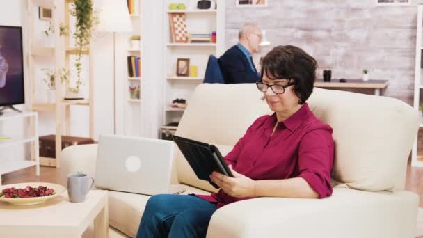 Ouderen leeftijd vrouw met behulp van tablet zitten op de bank in de woonkamer — Stockvideo