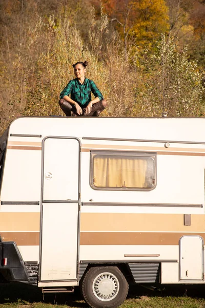 Chica joven sentada en el techo de una caravana retro y admirando la vista de otoño —  Fotos de Stock