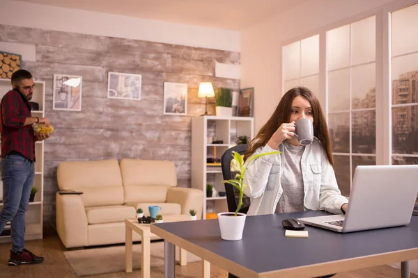 Freundin nimmt einen Schluck Kaffee, während sie im Wohnzimmer am Laptop arbeitet — Stockfoto