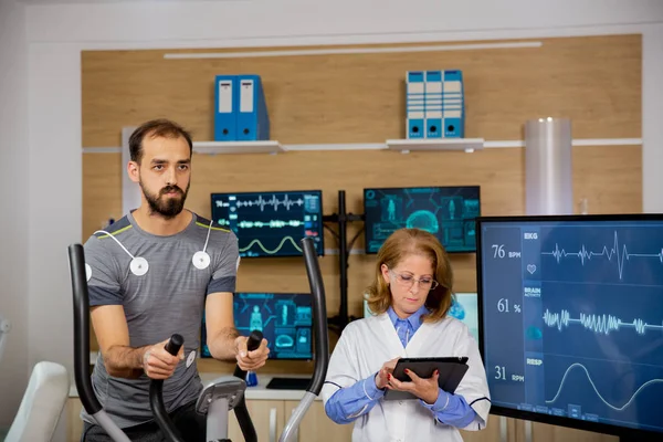 Manlig idrottsman som gör en fysisk ansträngning på trappan och har elektroder knutna till honom och läkaren som följer tabletten och antecknar — Stockfoto