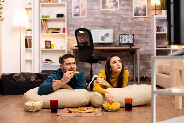 Пара, сидящая на полу и смотрящая телевизор в своей гостиной — стоковое фото