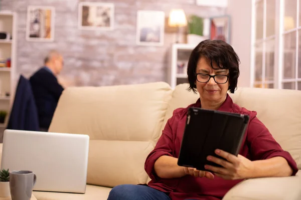 Oude oudere vrouw zittend op de bank en gebruikmakend van een digitale tablet Pc in gezellige woonkamer — Stockfoto