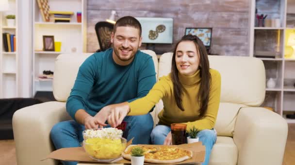 Pasangan duduk di sofa tertawa sambil menonton tv dan makan pizza — Stok Video
