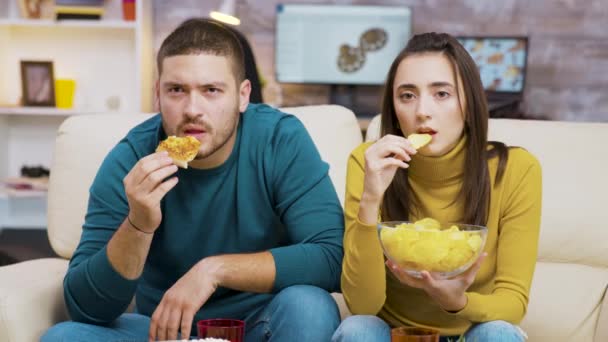 Przerażona para podczas oglądania telewizji i jedzenia niezdrowego jedzenia — Wideo stockowe