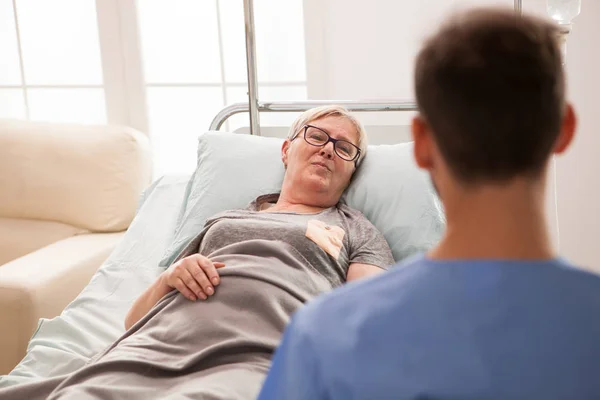 Vieja mujer acostada en la cama en un asilo de ancianos hablando con un médico —  Fotos de Stock