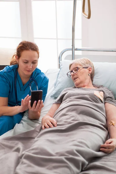 Vrouwelijke dokter helpt oude vrouw zitten op bed — Stockfoto