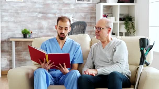 Maatschappelijk werker leest een boek voor aan een oude gehandicapte man in het verpleeghuis — Stockvideo