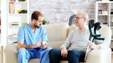 Huzurevinde erkek hemşire yanında koltuk değnekleriyle yaşlı bir adamla konuşuyor.