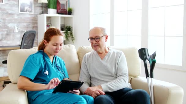女性の白人看護師は、明るく居心地の良い老人ホームで高齢者の健康についてメモを取る — ストック動画