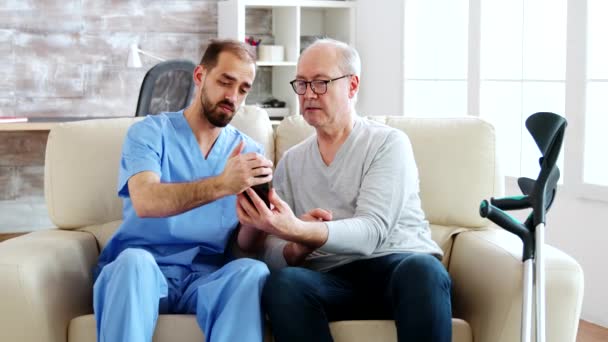 Mannelijke verpleegster helpt een gepensioneerde oude man om een smartphone te gebruiken — Stockvideo