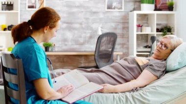 Hemşire ona kitap okurken, yaşlı bir kadının hastane yatağında yattığını gösteren bir fotoğraf.