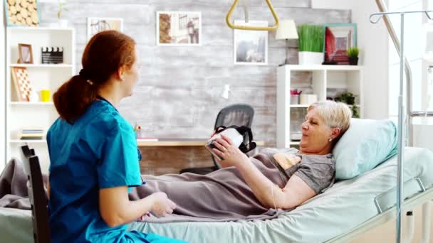 Enfermeira em casa de aposentadoria dando a uma senhora idosa com deficiência um fone de ouvido VR — Vídeo de Stock
