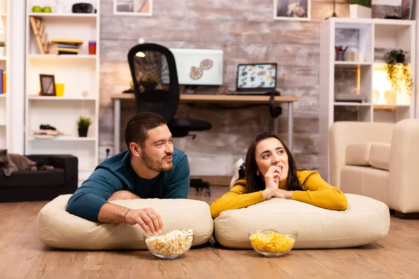 Пара, сидящая на полу и смотрящая телевизор в своей гостиной — стоковое фото