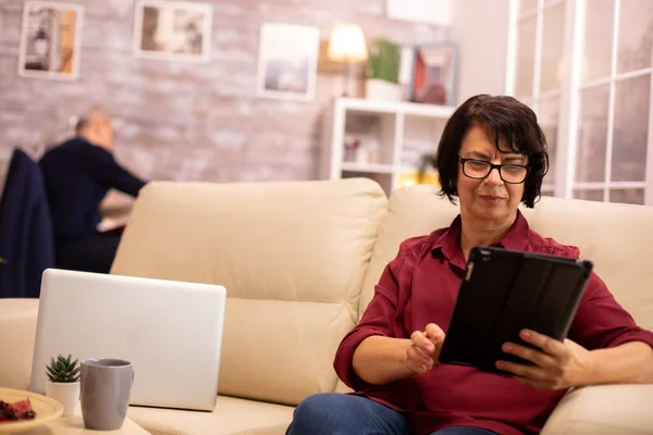 Oude oudere vrouw zittend op de bank en gebruikmakend van een digitale tablet Pc in gezellige woonkamer — Stockfoto