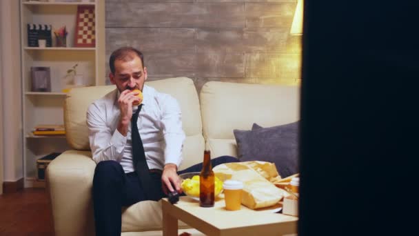 Бизнесмен с галстуком сидит на диване и ест бургер. — стоковое видео