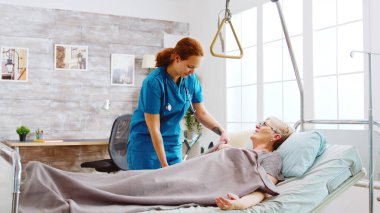Female nurse helping an old disabled and retired woman to go to bed clipart
