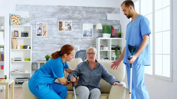 Femmina medico e il suo assistente aiutare la vecchia donna con stampelle a stare in piedi dal divano — Foto Stock