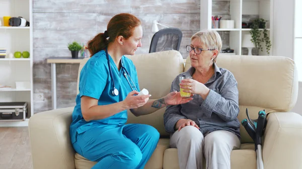 Medico femminile seduto sul divano con la donna anziana in casa di cura dando le sue pillole quotidiane — Foto Stock