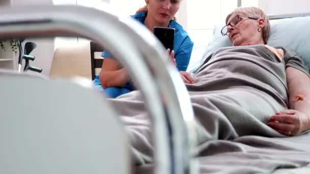 Женщина-медсестра помогает пожилой женщине использовать смартфон — стоковое видео